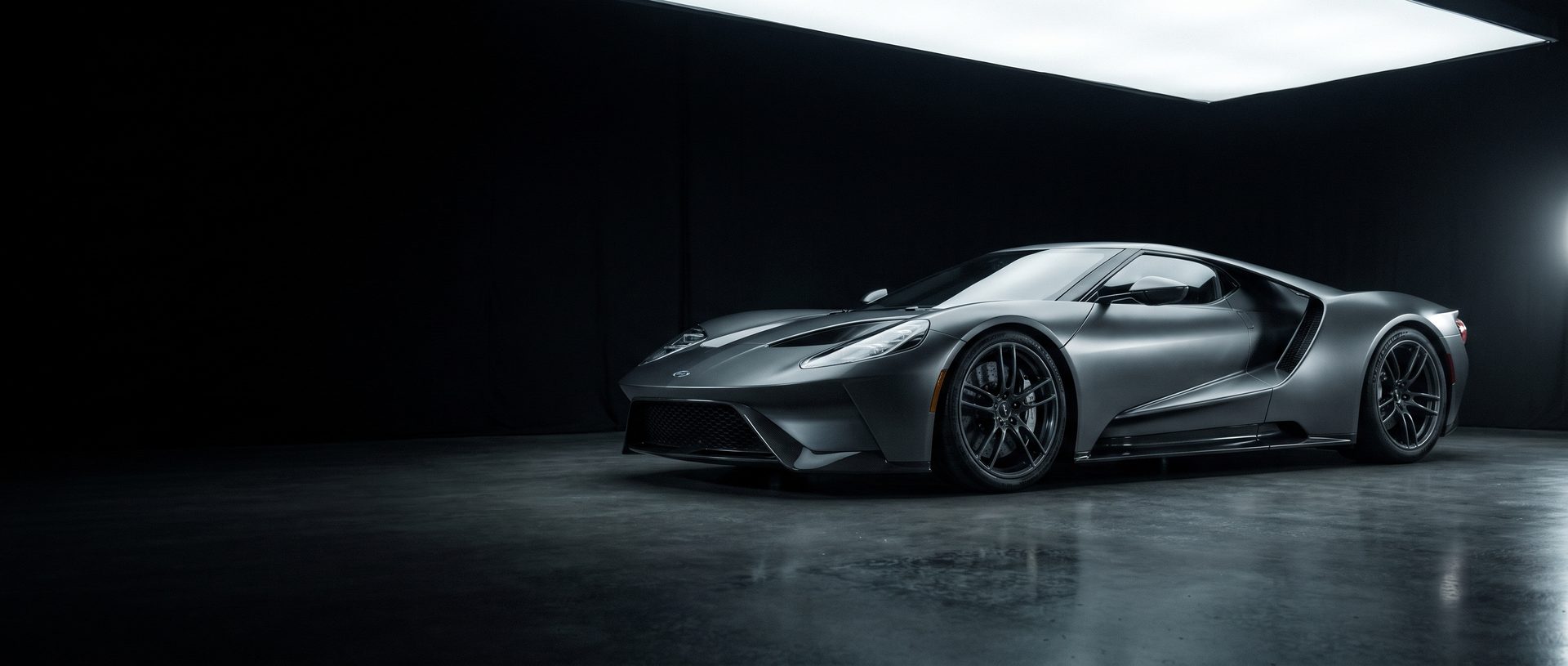 A satin graphite mid-engine supercar with a carbon front splitter and forged wheels, lit by a single overhead softbox on polished concrete